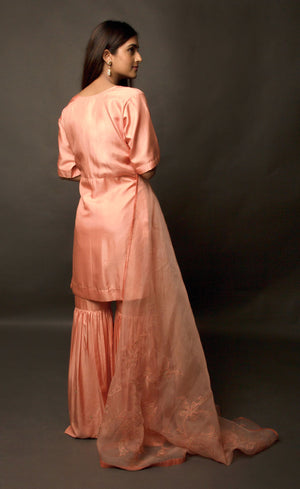 This is a beautiful burnt peach gharara suit set. The muse is wearing a simple yet an elegant kurta with yoke along the waistline paired with gharara having intricate  zari motifs on it. The suit-set is further accompanied by an organza dupatta with appliqué embroidery on it along the width and is finished with a raw silk border.