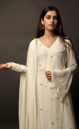 The muse is seen here wearing a classic angrakha style kurta suit set, delicately hand embroidered with a steel grey tilla. The motifs are scattered all over the front and back of this ivory angrakha set in georgette. The double bell sleeve with a beautiful flare, is a little modern play here in this classic angrakha silhouette. The silhouette is paired with wide legged easy palazzos and a dupatta embellished with tonal grey beadwork border.