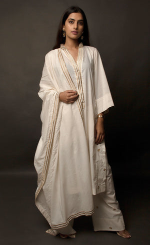 The muse is wearing a classic kurta set style, enhanced with added godet panels. This kurta is crafted in 100% cotton, is delicately embroidered with aari zari work paired with relaxed palazzos and accompanied by a 100% cotton dupatta embroidered with a coordinating zari border.