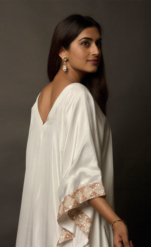 The muse is seen here wearing a classic V neck satin kaftan hand embellished with sequins and beads. The bordered embroidery beautifully runs along the length of the satin kaftan and also along its relaxed sleeves. The embellishments are all tonal shades of peach and off white.