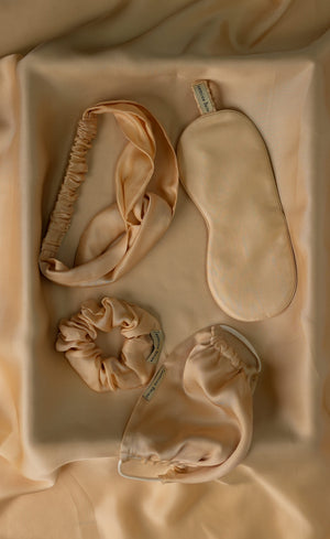Set of beige satin sleep accessories including a headband, eye mask, a scrunchy and a face mask on a beige fabric background.