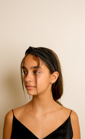 A model wearing a black satin single knotted headband in a studio. 