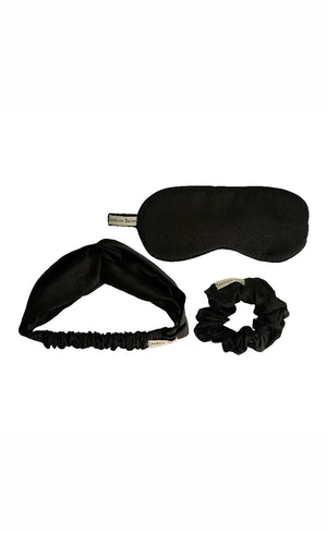 A black satin accessories set of eye mask, headband and a scrunchy on a flay lay background. 