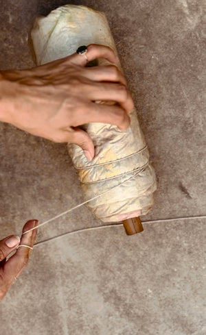 Hands tightly rolling and binding botanical-laden silk fabric around a wooden dowel with string -  the bundling step in the eco-printing process before steaming. 
