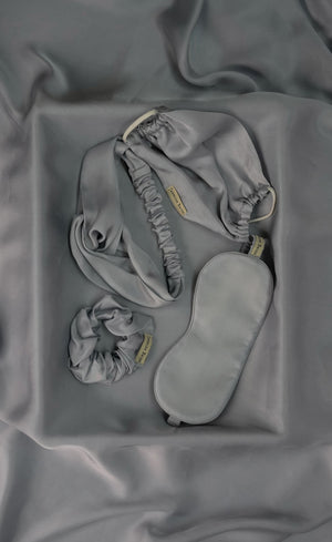 Set of gray sleepwear including a headband, eye mask, and scrunchie on a matching fabric background.