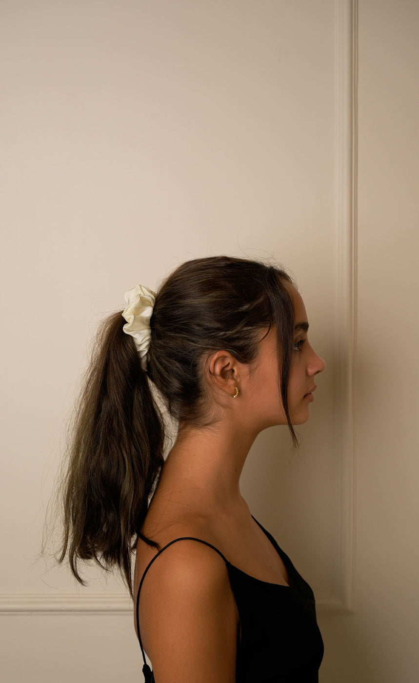 Woman with a ponytail wearing a white scrunchy against a neutral wall.