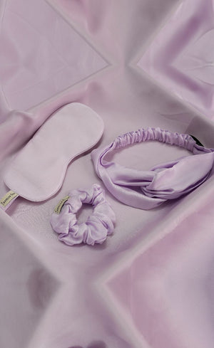 Set of lavender eye mask, headband, and scrunchie on a soft pink fabric background