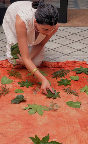 Jasmine Bains arranging fresh leaves - fig, fern, and tropical foliage -  on madder-orange eco-printed silk fabric, preparing for the botanical print process. 
