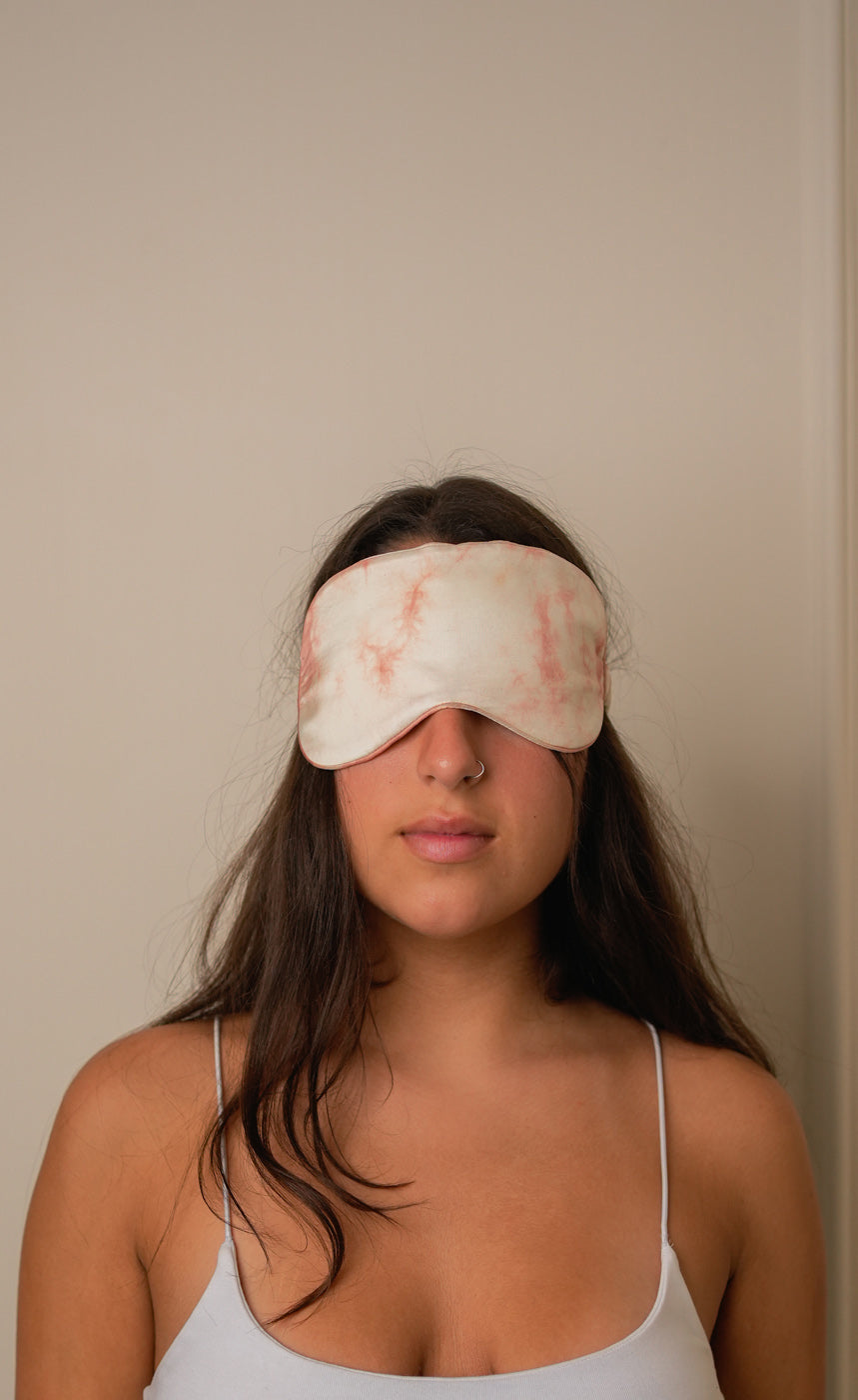 Woman wearing a light-colored sleep mask indoors against a plain wall.