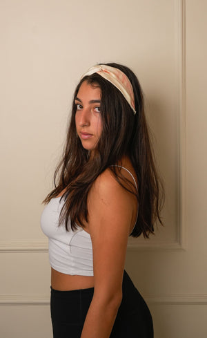 Woman wearing a tie and dye rust beige knotted headband against a beige wall.
