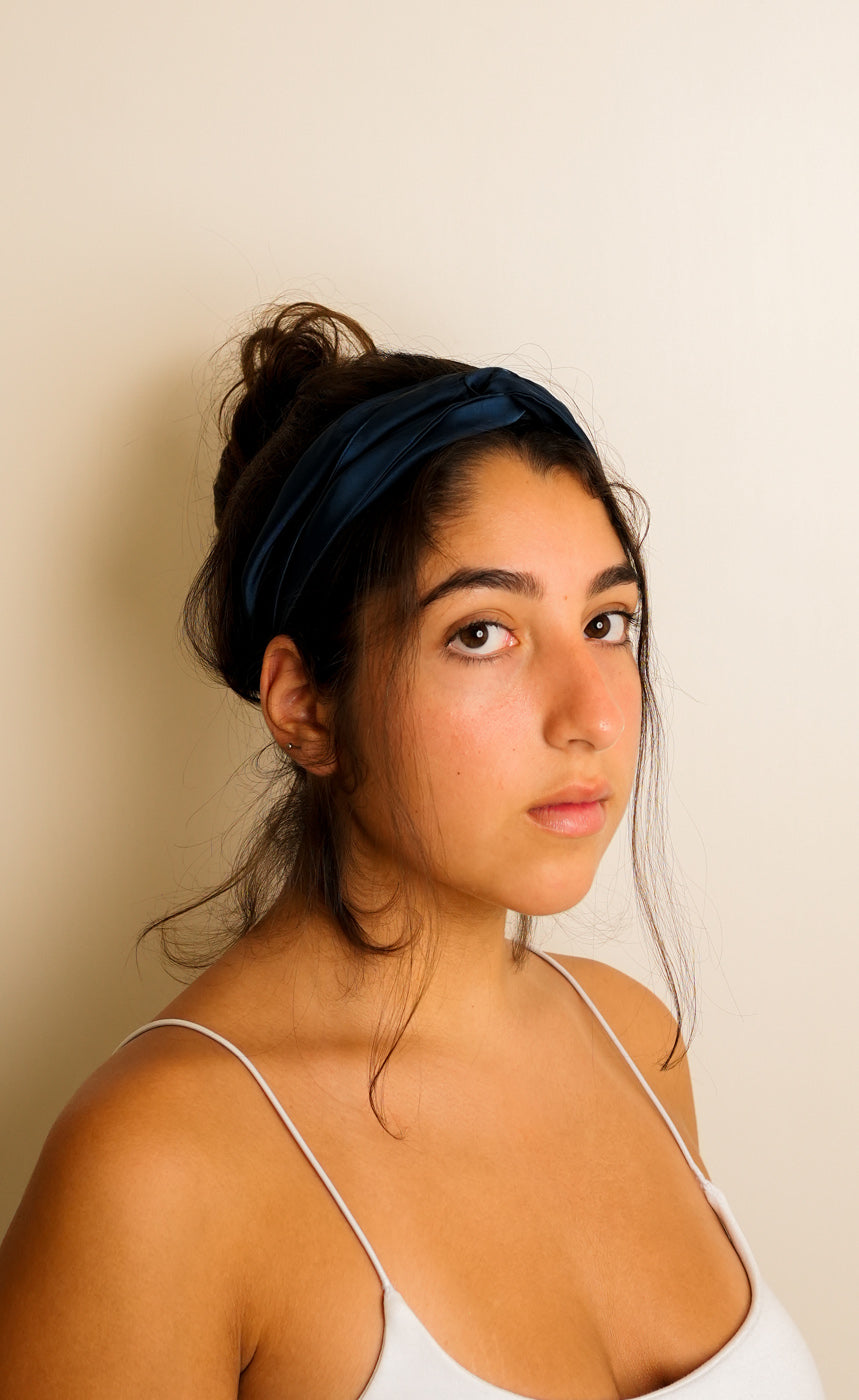 Woman wearing a blue knotted satin headband against a light coloured wall. 