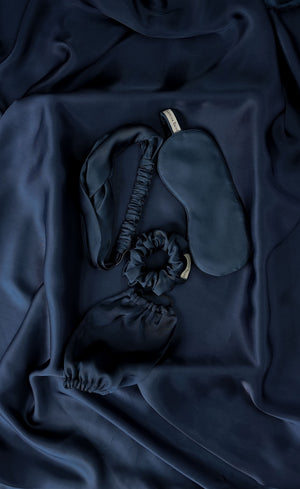 Set of dark blue satin eye mask, headband, and a scrunchie on a dark fabric background