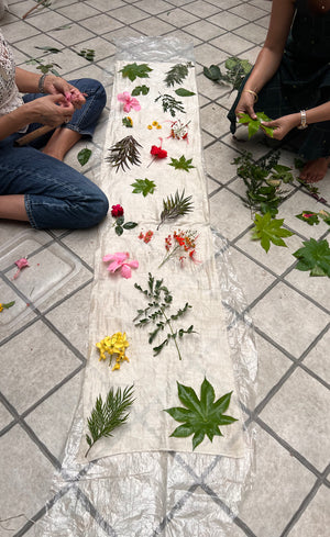 Workshop participants arranging a pattern of fresh botanicals - hibiscus, pink hibiscus, silver oaks, castor, rose leaves and flowers, amaltas flowers and peacock flowers  along a length of silk fabric scarf  laid on the floor, designing the composition before eco-printing. 