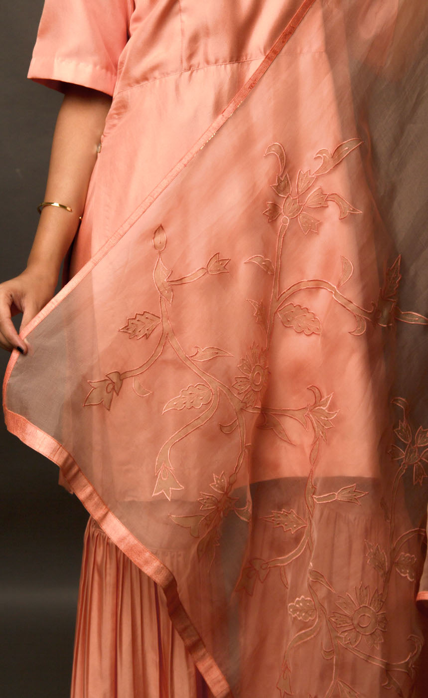 This is a beautiful burnt peach gharara suit set. The muse is wearing a simple yet an elegant kurta with yoke along the waistline paired with gharara having intricate  zari motifs on it. The suit-set is further accompanied by an organza dupatta with appliqué embroidery on it along the width and is finished with a raw silk border.