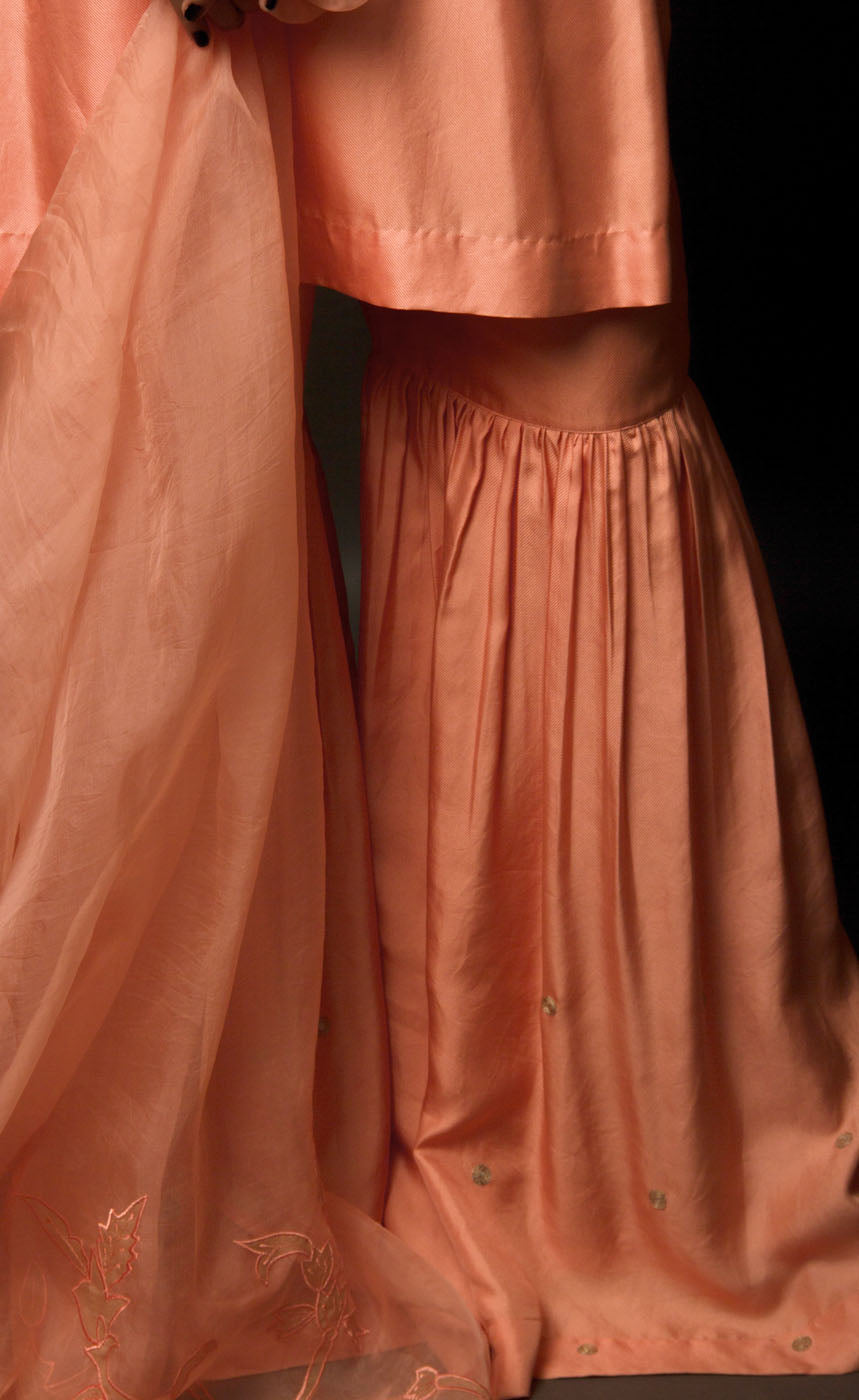 This is a beautiful burnt peach gharara suit set. The muse is wearing a simple yet an elegant kurta with yoke along the waistline paired with gharara having intricate  zari motifs on it. The suit-set is further accompanied by an organza dupatta with appliqué embroidery on it along the width and is finished with a raw silk border.