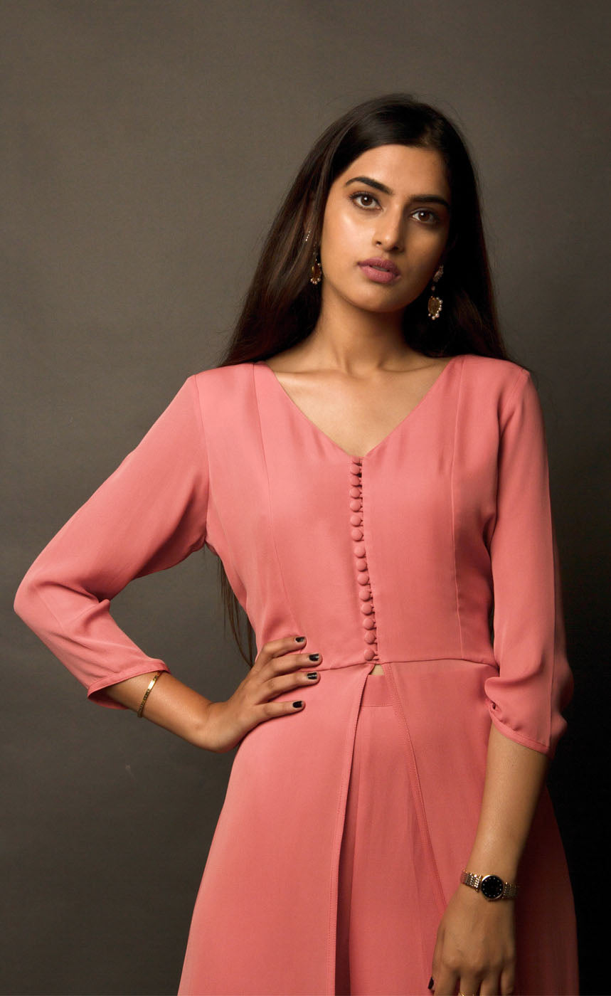 The muse here is wearing a simple and elegant front-slit dress with palazzos in carrot pink. The dress comprises of an upper yoke and a simple v-neckline which is further having a vertical buttoned detail on yoke and a high slit in the middle center. It is further paired with cupro georgette flowy wide legged palazzos.