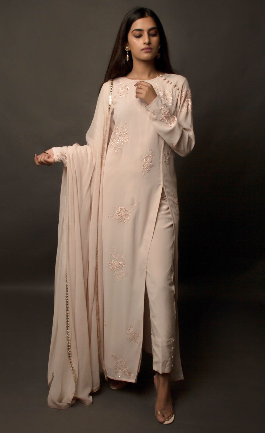 Dusty Rose side slit suit set, crafted in georgette, fully embroidered with signature flat threadwork topped with sequins spray accompanied by a dupatta with rose gold mukaish border. The ensemble carries a strong of a confident style because of the slim straight trousers underneath, the bottom of the trousers is finished by our signature buttoned hemline style. Flaunt your chic style in this elaborately worked ensemble, the muse radiates a modern, yet classic vibe. 