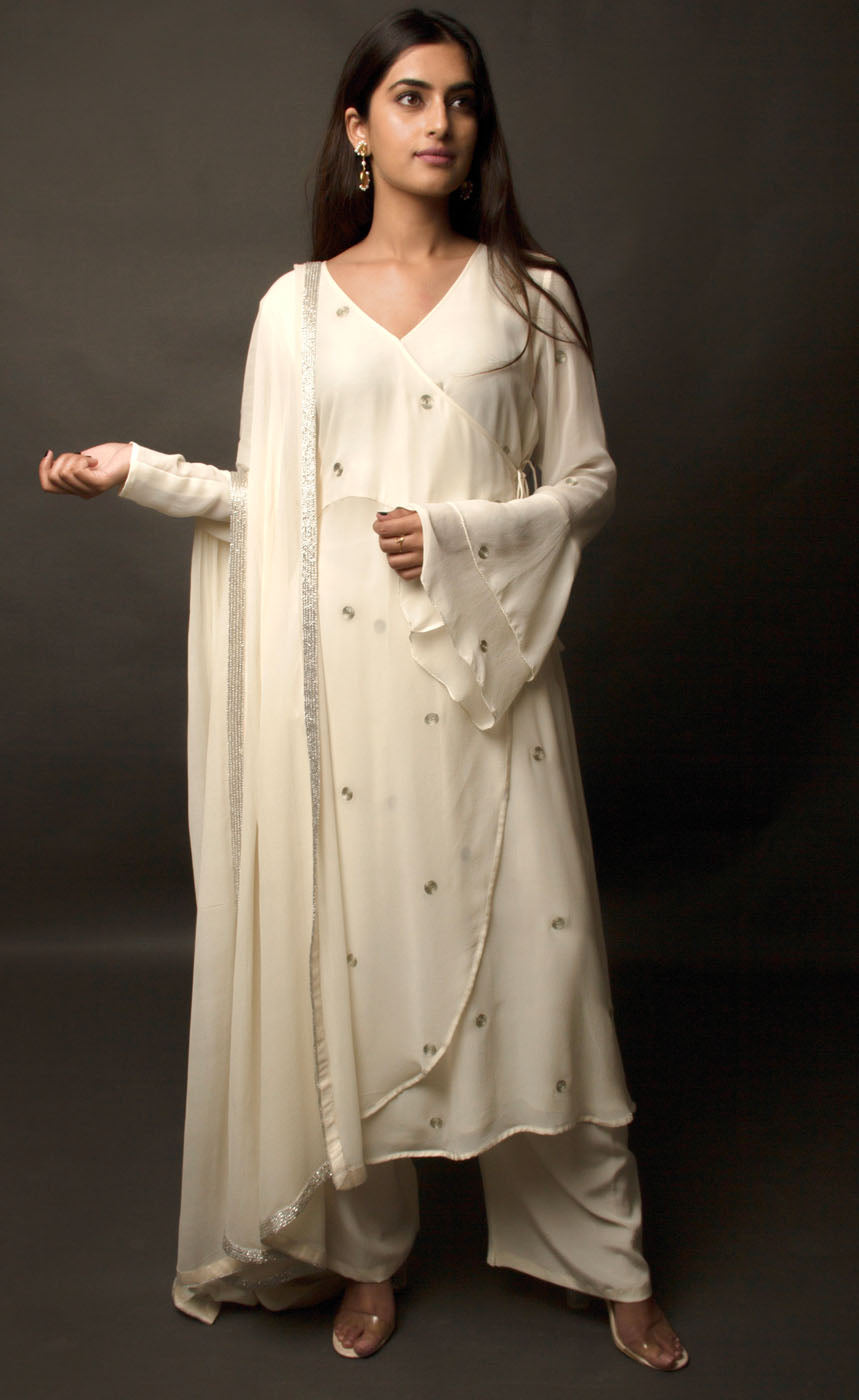 The muse is seen here wearing a classic angrakha style kurta suit set, delicately hand embroidered with a steel grey tilla. The motifs are scattered all over the front and back of this ivory angrakha set in georgette. The double bell sleeve with a beautiful flare, is a little modern play here in this classic angrakha silhouette. The silhouette is paired with wide legged easy palazzos and a dupatta embellished with tonal grey beadwork border. 