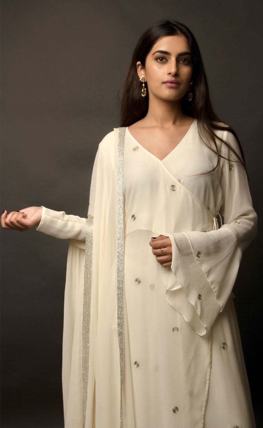 The muse is seen here wearing a classic angrakha style kurta suit set, delicately hand embroidered with a steel grey tilla. The motifs are scattered all over the front and back of this ivory angrakha set in georgette. The double bell sleeve with a beautiful flare, is a little modern play here in this classic angrakha silhouette. The silhouette is paired with wide legged easy palazzos and a dupatta embellished with tonal grey beadwork border. 