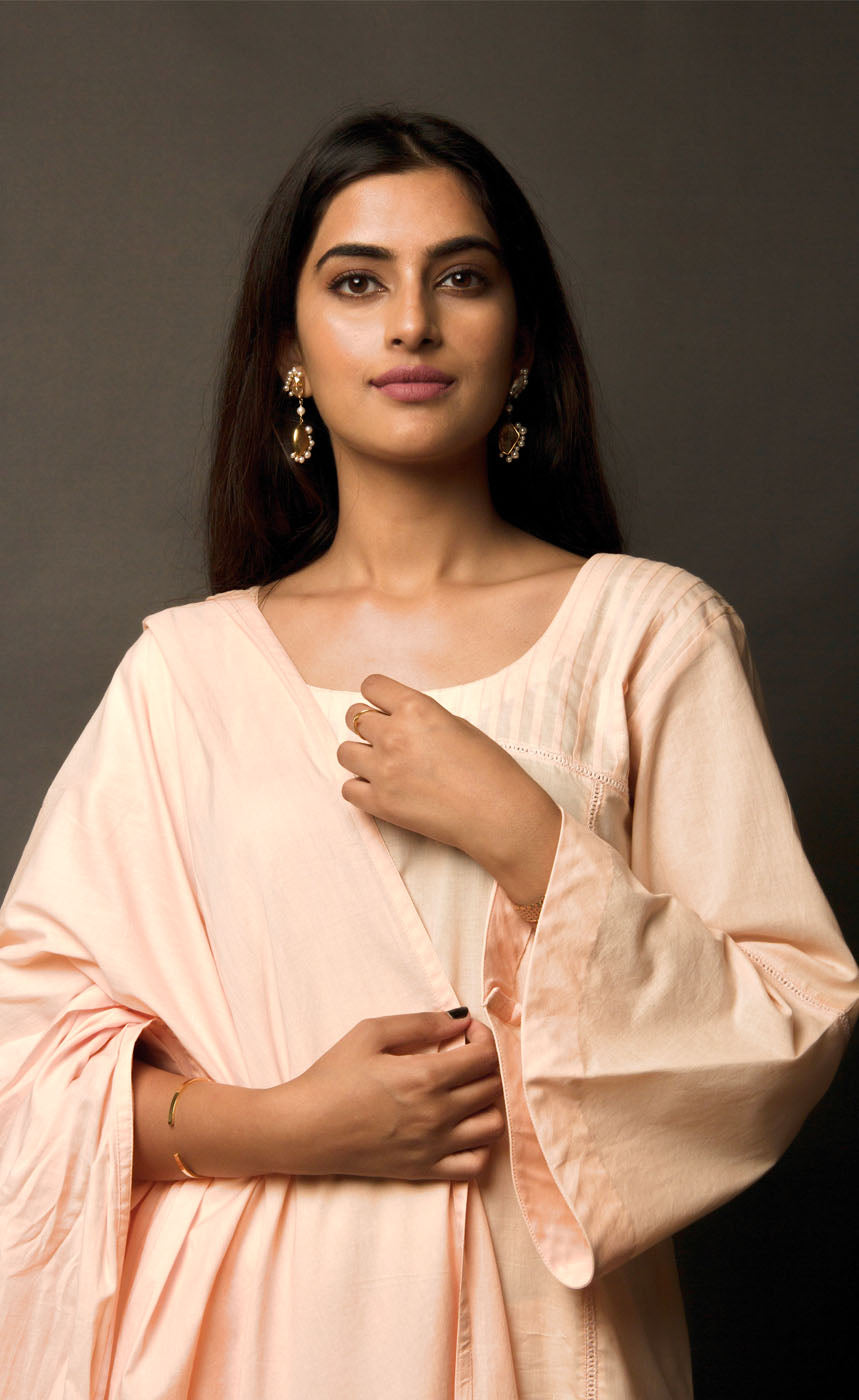 The muse here is wearing a peach embroidered kurta suit set. This is a 100% cotton suit with panelled kurta having lace insertions in the vertical panels and pin tucks in the upper yolk. The kurta is enhanced with flared sleeves having single horizontal lace insertion around the elbow. It is paired with a flared cotton palazzos and a cotton dupatta with our signature thread-work embroidery along its width, further finished with a buttoned detail. 