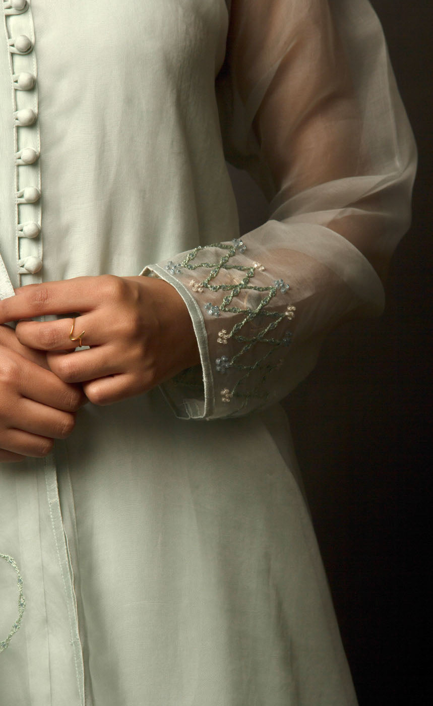 The muse here is wearing a pista green silk organza suit set intricately embroidered with our signature thread-work. The look comprises of sheer silk organza kurta having a detailed buttoned neckline with bead-work embroidery on the sleeves and back terrace, further lined with a cupro crepe slip. It is paired with an organza dupatta embroidered along the length and is further finished with raw silk and crepe pants. 