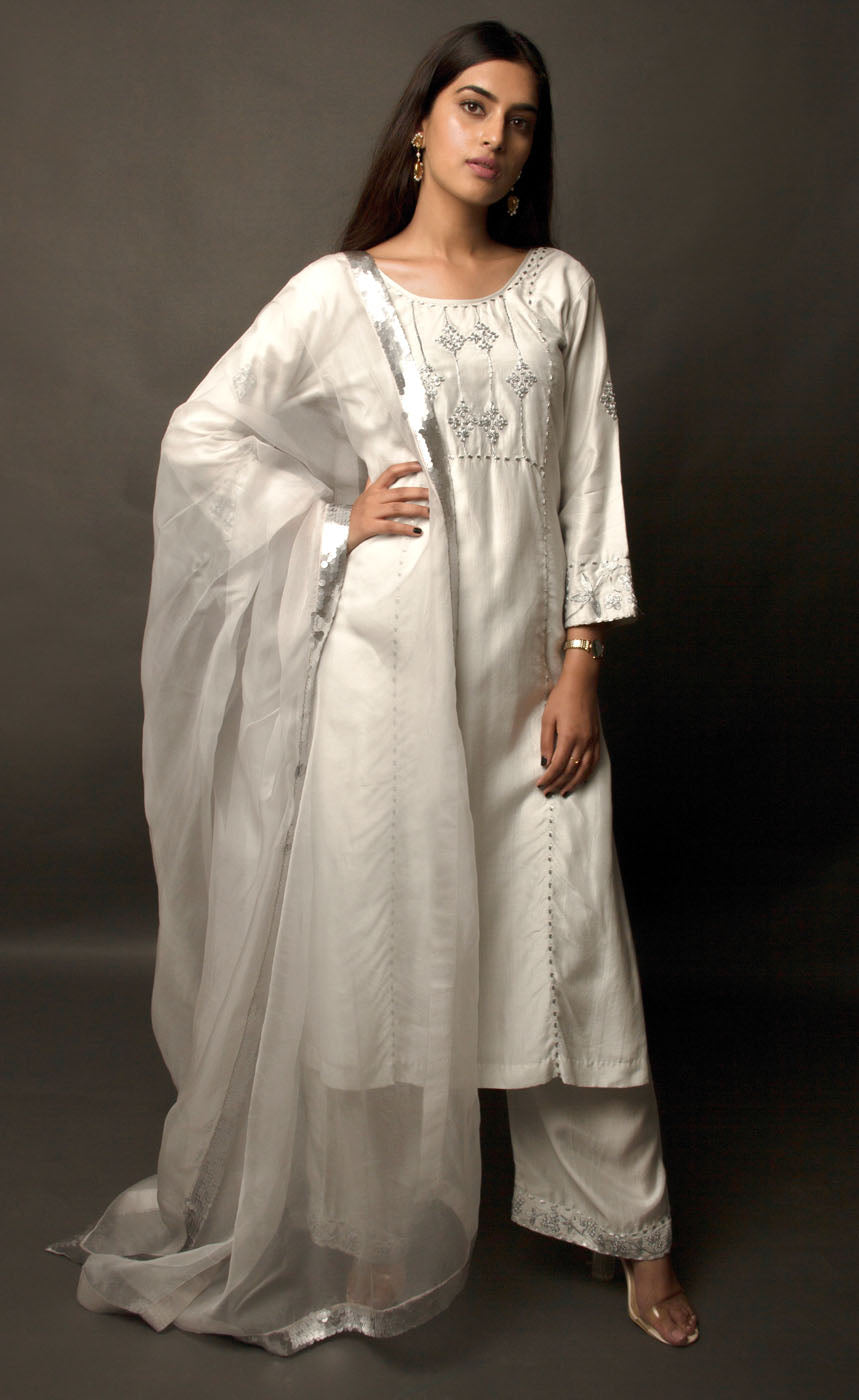 The muse is wearing a classic powder grey intricately embroidered kurta suit set with palazzos. The yoke panel, along with sleeve cuff and the palazzos bottom/cuff are delicately embroidered with silver-grey tilla. It is completed with a sheer organza dupatta carrying a matte grey hand-embellished sequin border.