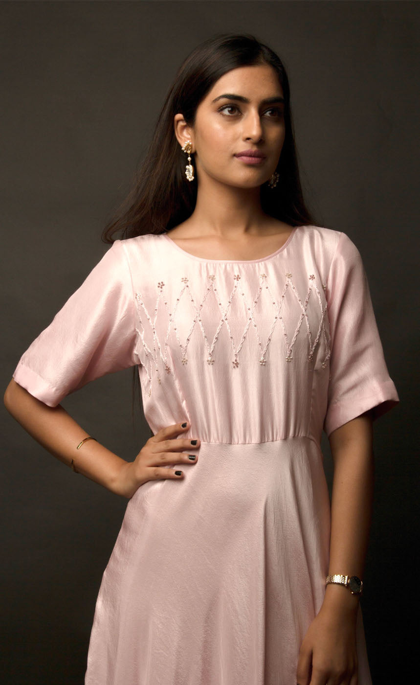 Graceful, demure and classic simplicity, this powder pink ensemble features a geometric hand embroidery on the mid yoke of this floor length flowing gown. Adorned with understated combination of peachy and translucent beads and threads, this cupro satin piece is yet a delicately detailed silhouette. 