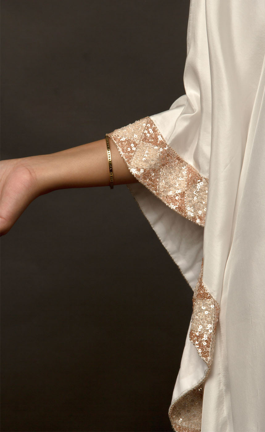 The muse is seen here wearing a classic V neck satin kaftan hand embellished with sequins and beads. The bordered embroidery beautifully runs along the length of the satin kaftan and also along its relaxed sleeves. The embellishments are all tonal shades of peach and off white. 